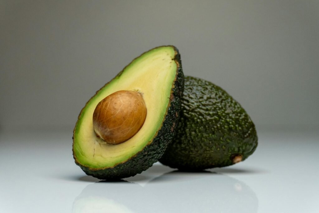 A cut avocado half with citrus juice and plastic wrap pressed directly onto the surface beside a small airtight container.